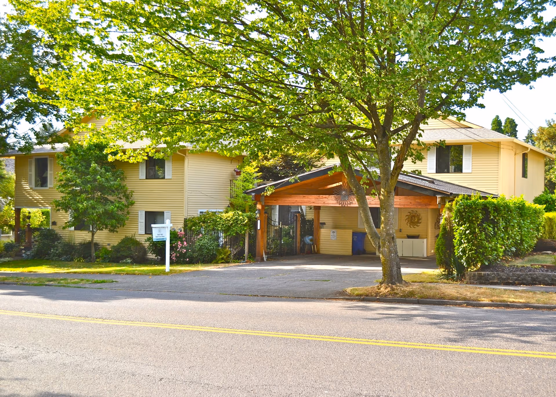 Exterior view of a two-story yellow building with white window shutters, surrounded by green trees and bushes. There is a covered driveway area with wooden beams and a paved road in the foreground.