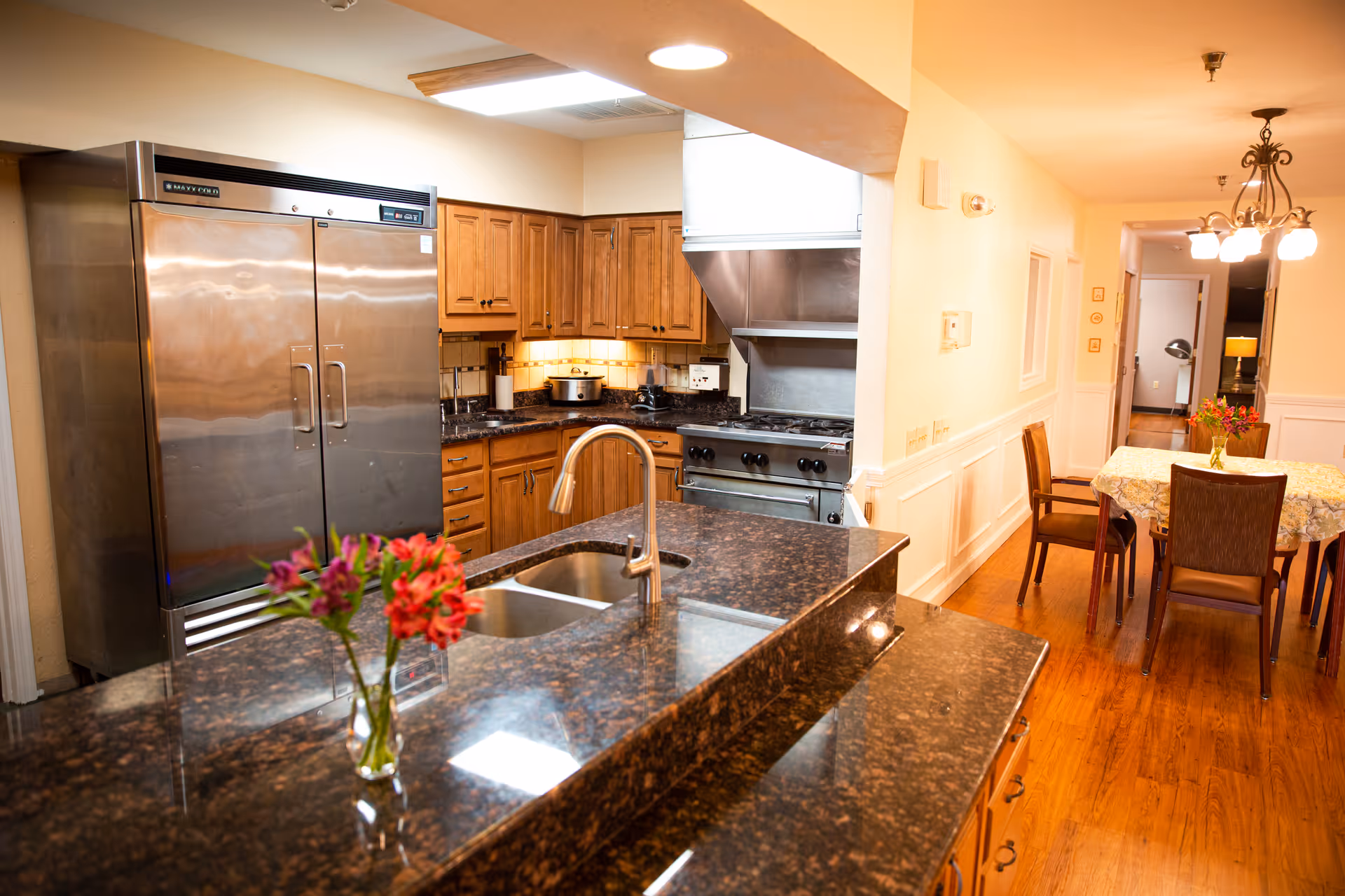 A kitchen area with a large stainless steel refrigerator, wooden cabinets, a stove with a vent hood, and a granite countertop with a sink and a vase of flowers. In the background, there is a dining area with a table covered with a floral tablecloth and four chairs, illuminated by a chandelier.