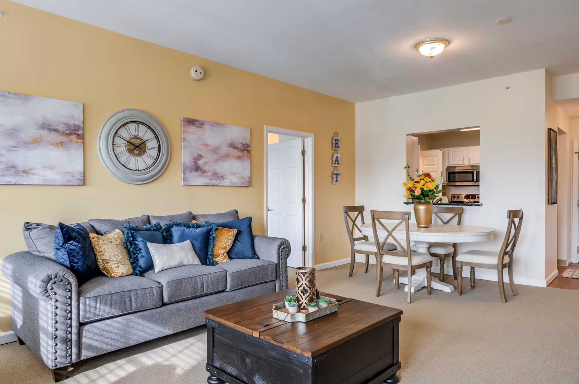A cozy living room and dining area with a gray sofa adorned with blue and gold pillows, a wooden coffee table with decorative items, a round dining table with four chairs, and a kitchen visible through a pass-through window. The walls are painted yellow and white, with abstract paintings and a large clock on the wall above the sofa.
