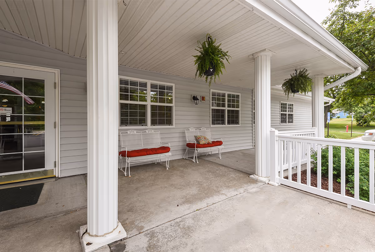Covered porch area with white pillars and railing at Amelia Senior Living. Two white metal benches with red cushions are placed against the wall under windows. Hanging green plants are suspended from the ceiling. A glass door entrance is visible on the left side, and greenery and trees are seen outside.