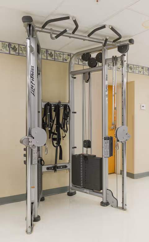 A Life Fitness multi-station cable machine in a room with beige walls and a floral border near the ceiling. Several black resistance straps hang on the wall behind the machine. The floor is light-colored, and a wooden door is visible in the background.