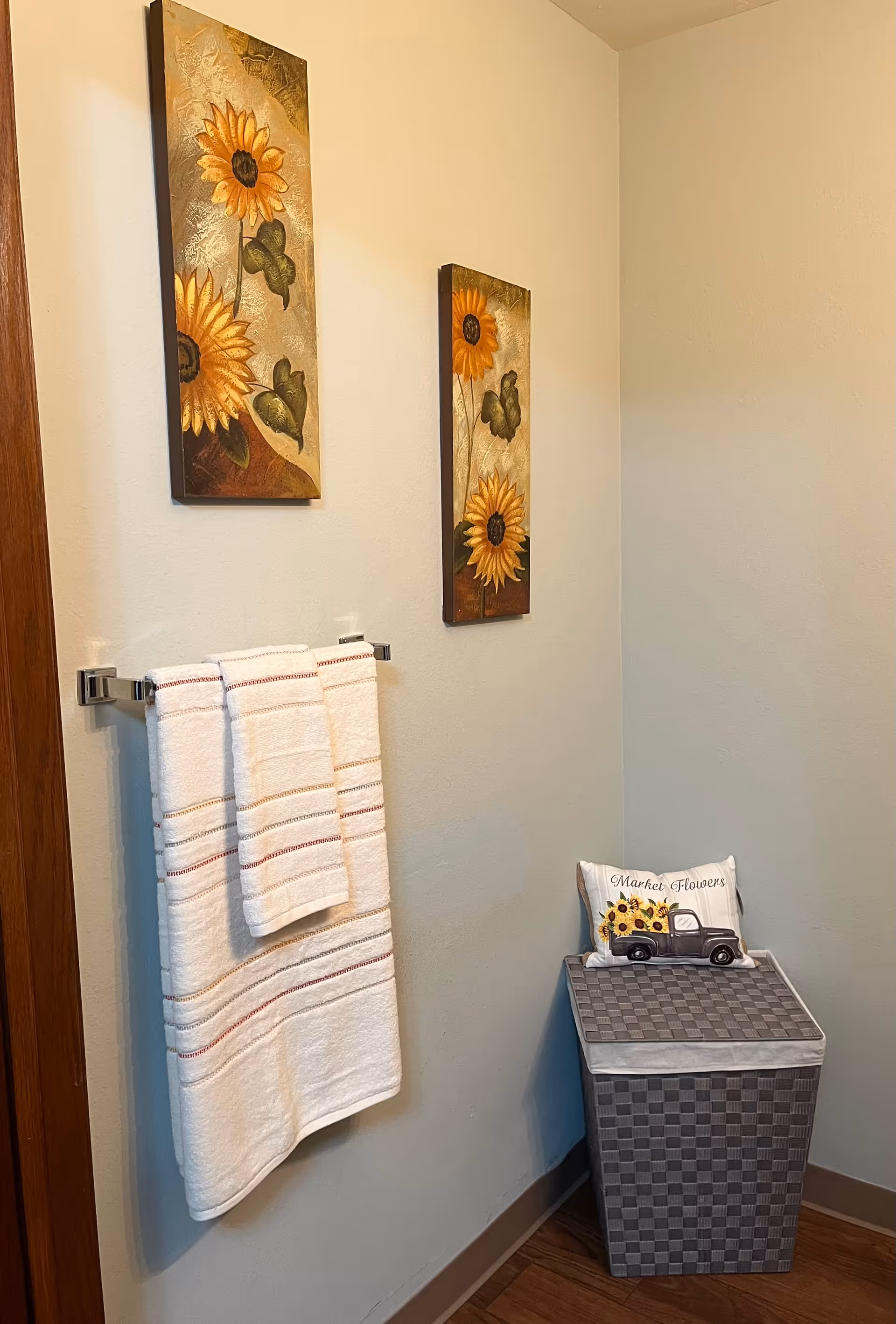 A corner of a bathroom with two sunflower-themed paintings on the wall, a towel rack holding three white towels with red and gray stripes, and a gray woven laundry hamper with a decorative pillow featuring a vintage truck and sunflowers that says 'Market Flowers'.