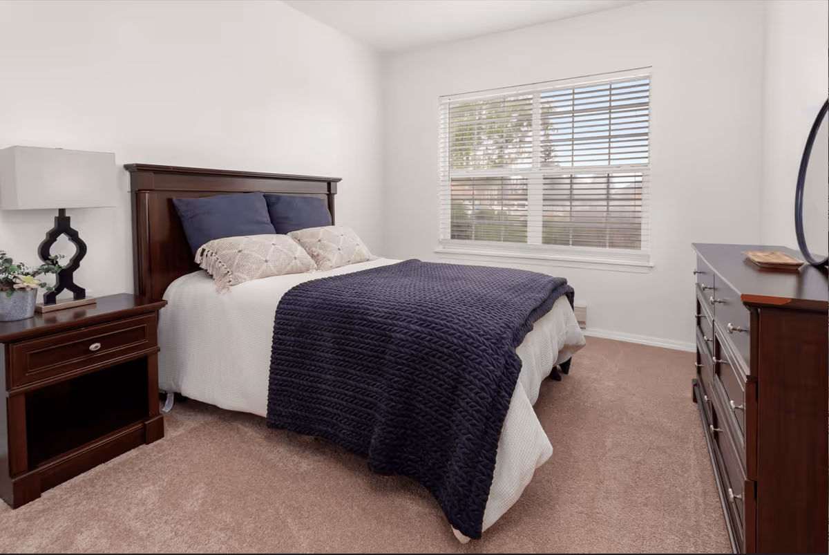 A bedroom with a neatly made bed featuring a dark wooden headboard, white bedding, and a dark blue knitted blanket. There are two beige decorative pillows and two dark blue pillows on the bed. To the left of the bed is a dark wooden nightstand with a modern lamp and a small plant. To the right is a matching dark wooden dresser with a round mirror on top. A window with white blinds lets in natural light, and the room has beige carpet and white walls.