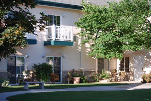 Exterior view of a senior living facility showing a beige two-story building with white balconies and large windows. There is a well-maintained green lawn with a stone bench, various potted plants, and outdoor seating areas. Trees provide shade and add to the landscaped garden setting.