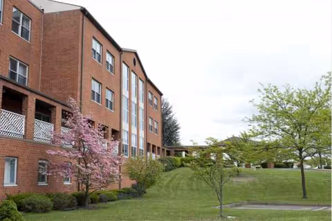 Red brick senior living building with green lawn, trees, and a pink flowering tree.