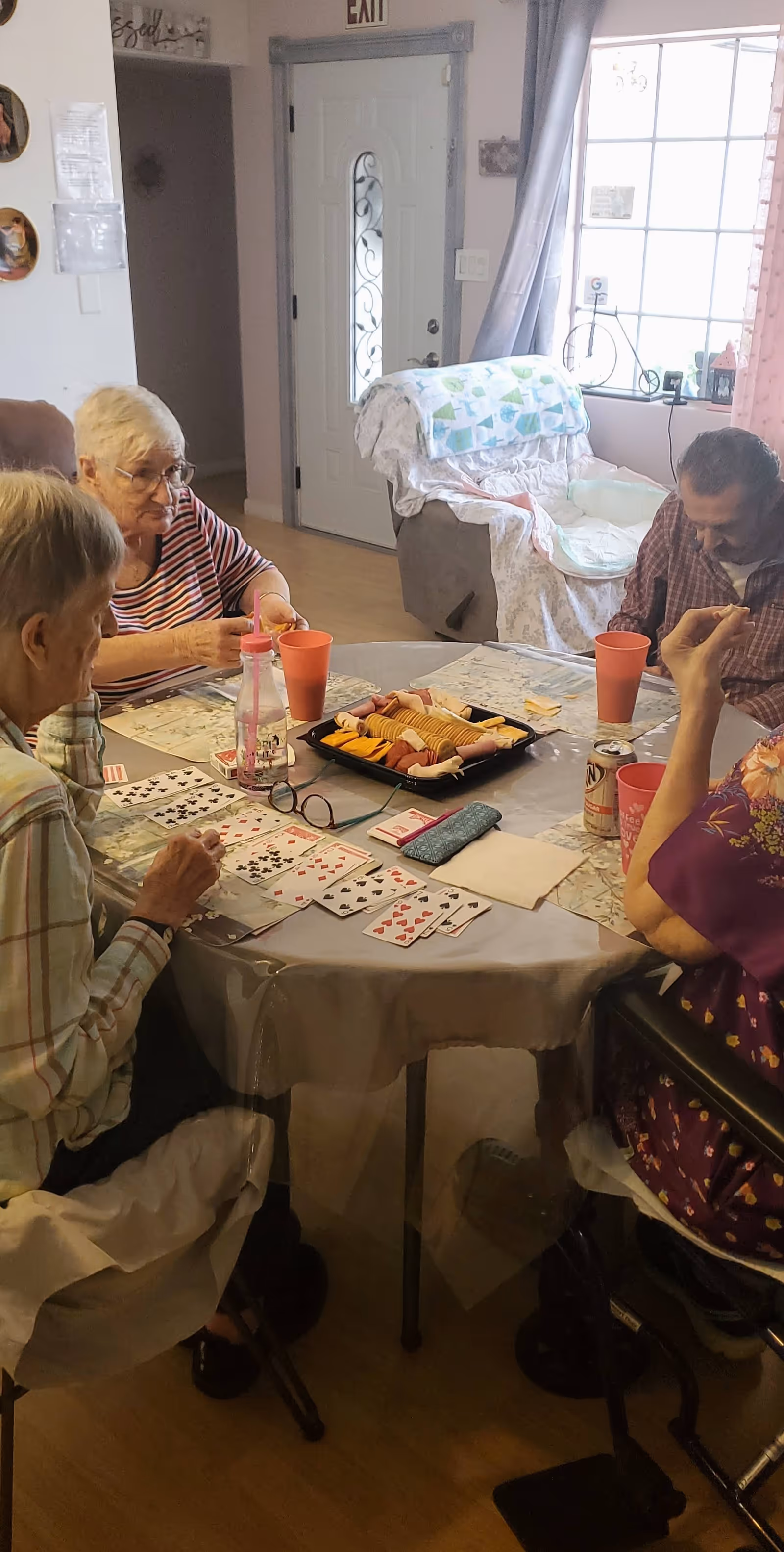 Four elderly people sitting around a round table playing cards in a cozy room with snacks and drinks on the table. There is a door and a window with curtains in the background, along with a cushioned chair covered with a blanket.