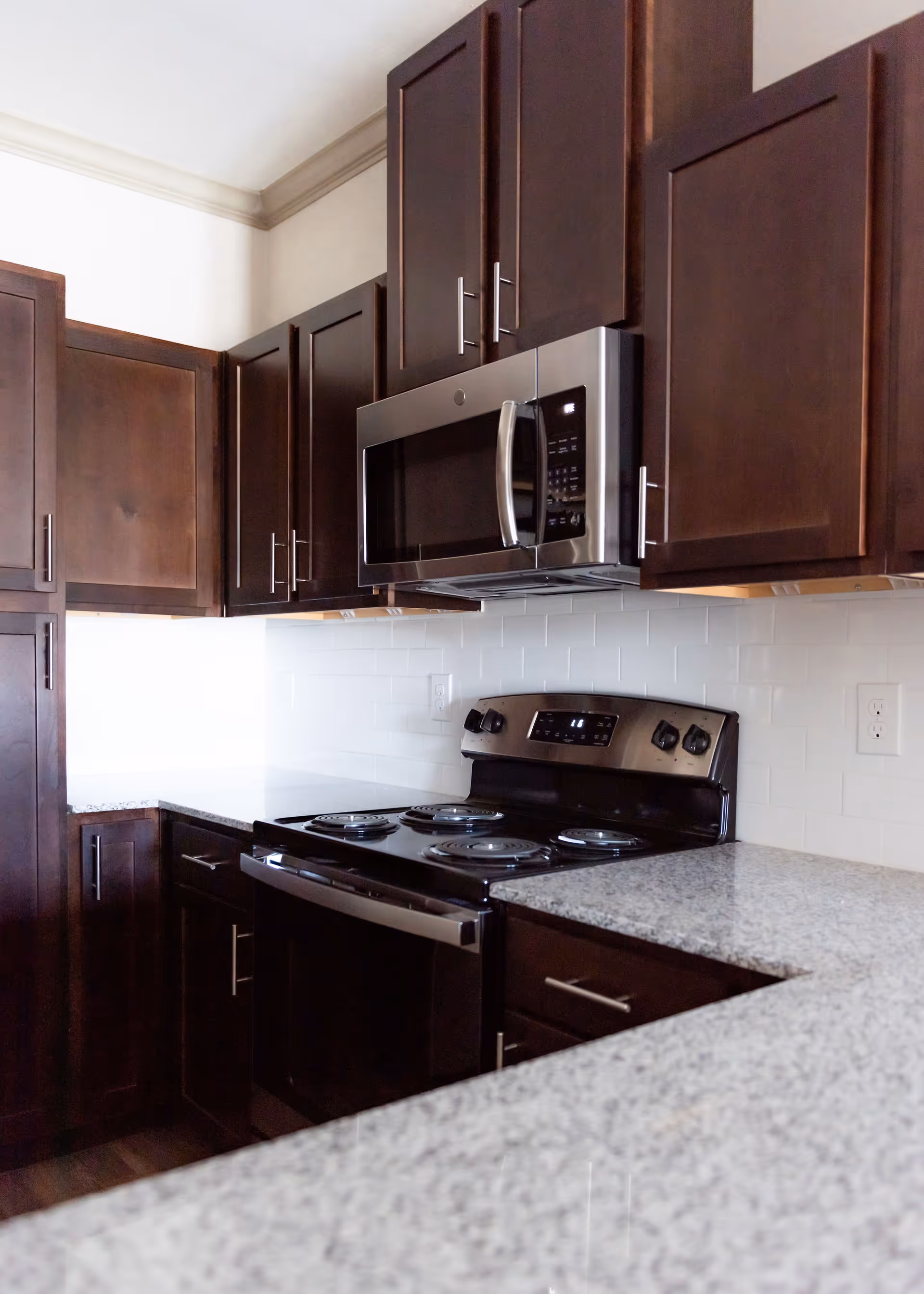 Modern kitchen with dark wooden cabinets, a stainless steel microwave mounted above a black electric stove, and light-colored granite countertops.