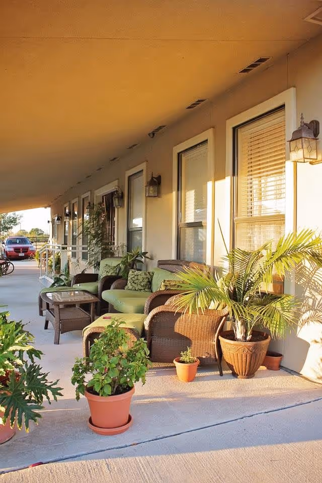 Covered outdoor patio area with wicker furniture including chairs and a loveseat with green cushions, a glass-top coffee table, and several potted plants. The patio is adjacent to a building with windows and wall-mounted lantern-style lights. A red car is visible parked in the background.