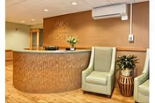 Reception area of Westgate Hills Rehabilitation & Nursing Center featuring a curved wooden reception desk with a flower arrangement, two green upholstered chairs, a small wooden side table with a plant, and a wall with the facility's name and logo.