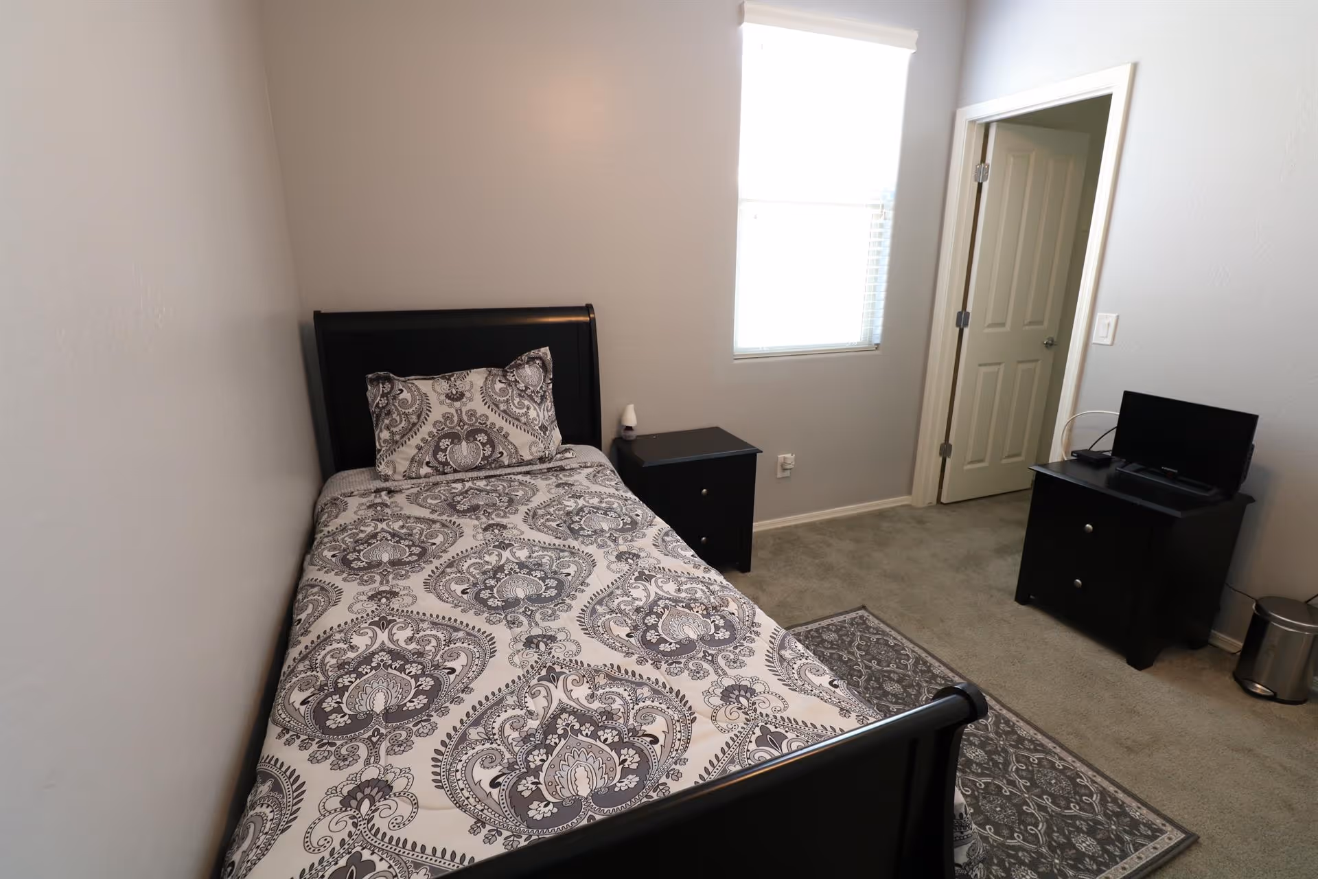 A small bedroom with a single bed featuring a black frame and patterned bedding in shades of gray and white. Next to the bed is a black nightstand with a small white object on it. There is a window with blinds on the wall opposite the bed, and an open door leading to another room. A black dresser with a small TV on top is positioned against the wall near the door. The floor is carpeted in gray, and a patterned rug lies beside the bed.