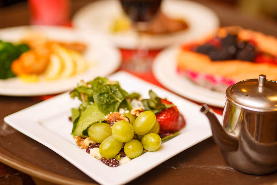 A close-up of a white square plate with a fresh salad consisting of green grapes, leafy greens, walnuts, and a strawberry. In the background, there are blurred plates of food and a metal teapot on the right side.