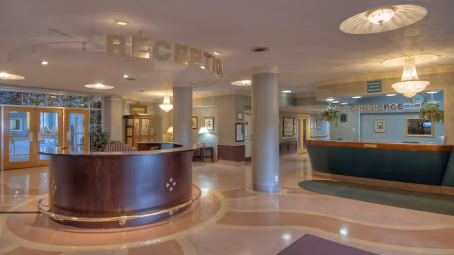 Interior view of a senior living facility lobby with a curved wooden reception desk on the left under a hanging sign that reads 'RECEPTION' and a concierge desk on the right under a sign that reads 'CONCIERGE'. The floor has a patterned design, and there are chandeliers and plants decorating the space.