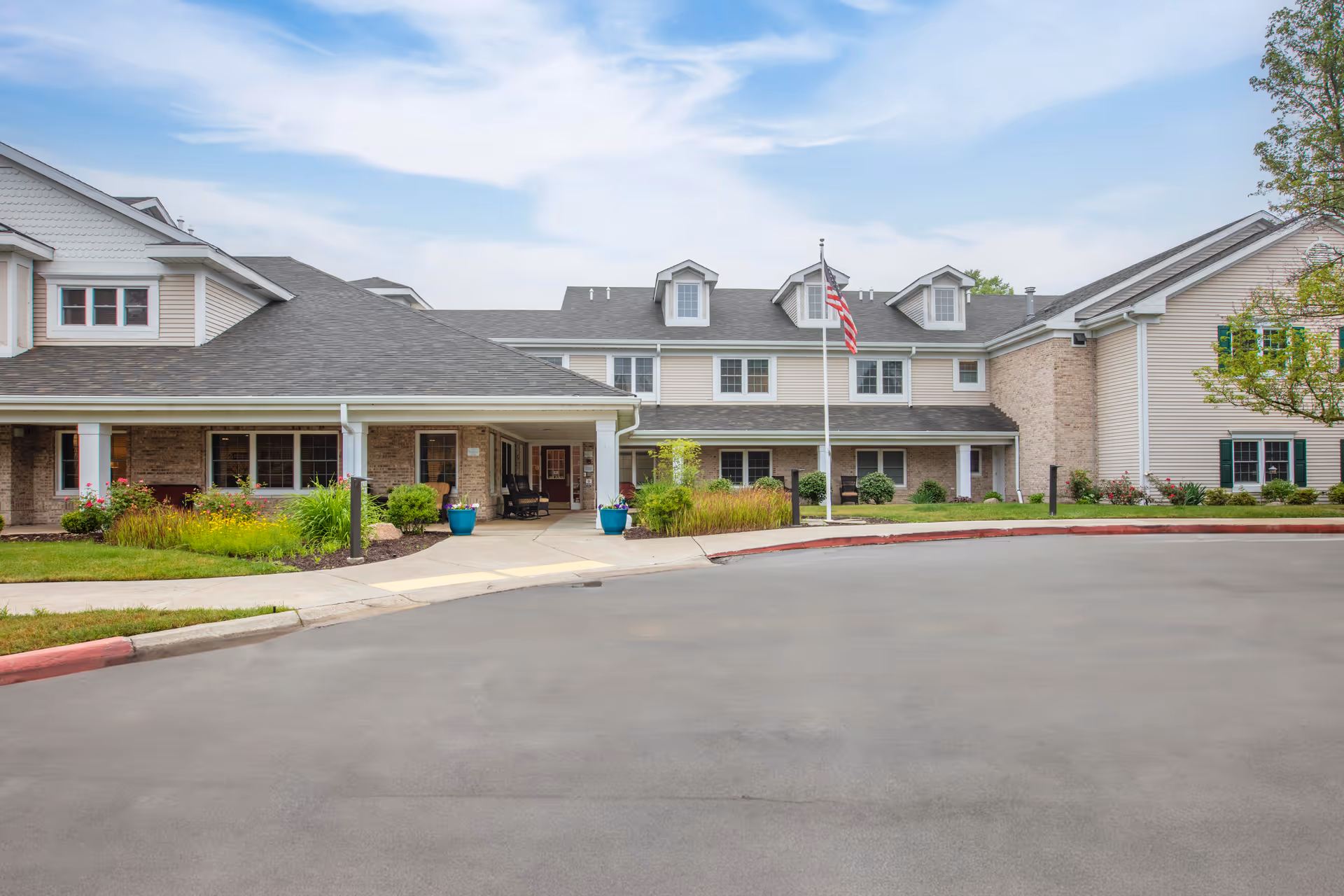 Exterior view of Brookdale Meridian - Assisted Living facility showing a two-story building with beige siding and brick accents, a covered entrance with seating, landscaped greenery, and an American flag on a flagpole in front.
