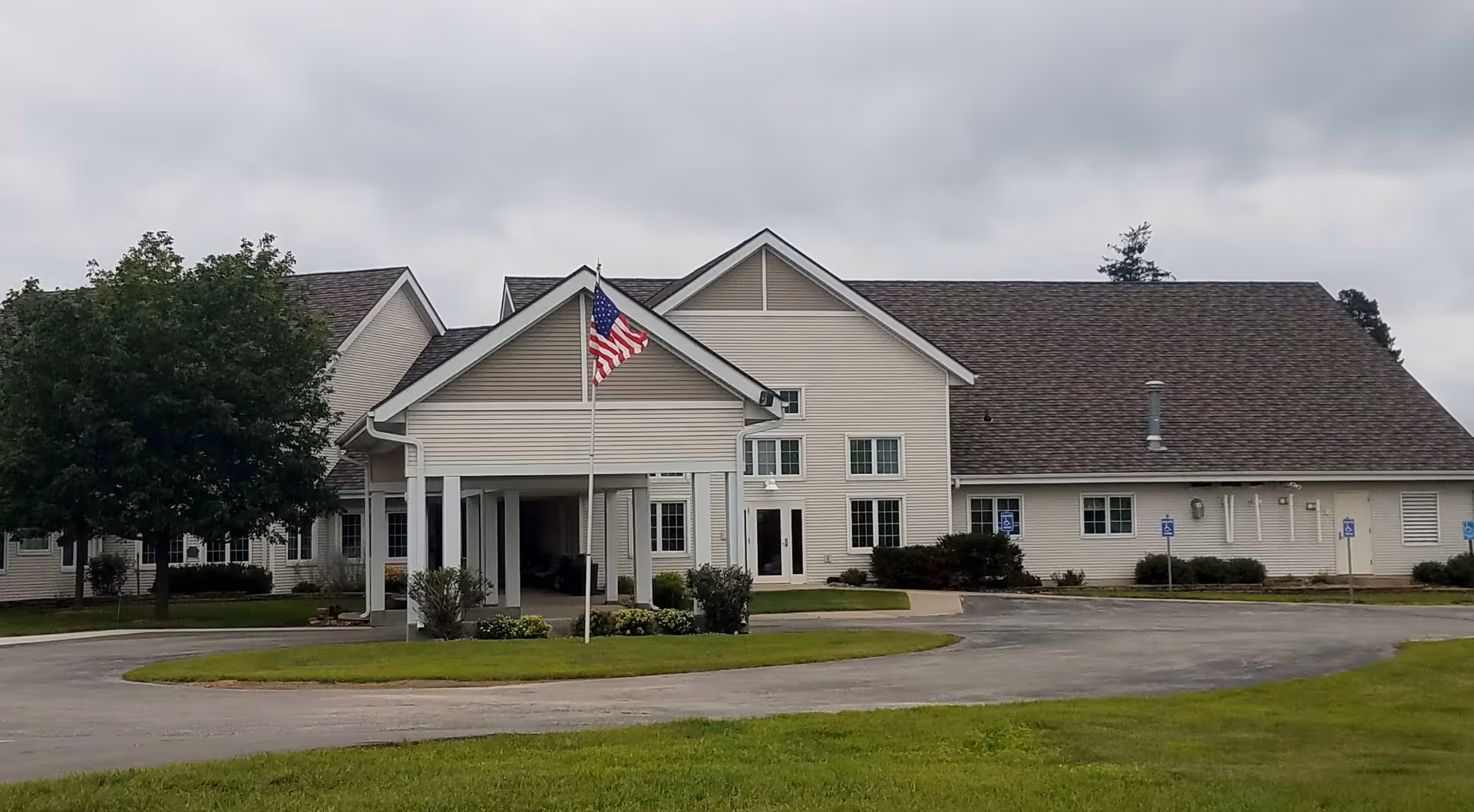 Exterior view of Hubbard Care Center, a single-story building with beige siding and a brown shingled roof. There is a covered entrance with white pillars and an American flag on a flagpole in front. The surrounding area includes a paved driveway, green grass, and a tree on the left side under a cloudy sky.