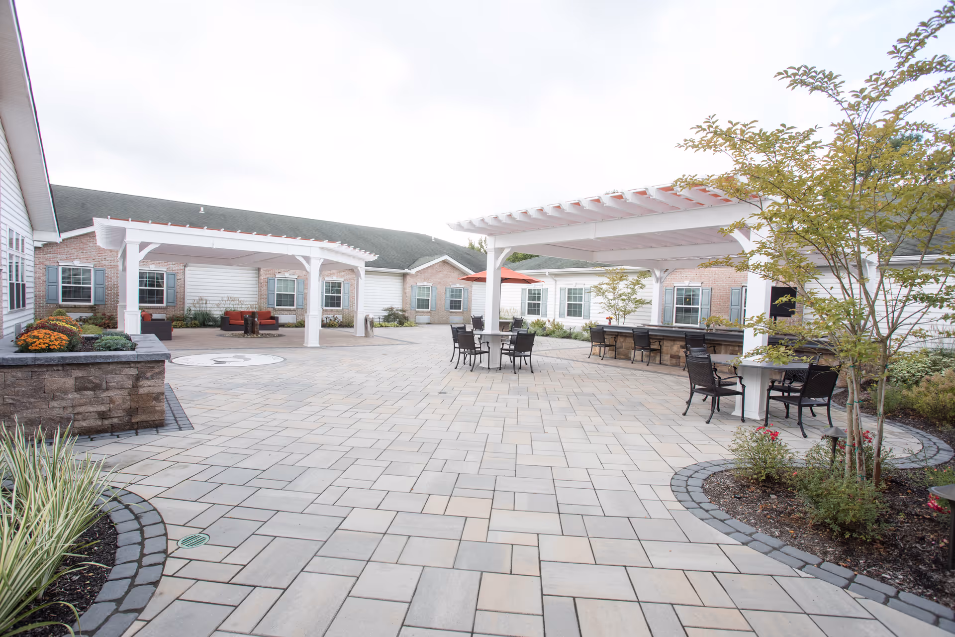 Outdoor courtyard area of CareOne at Evesham Assisted Living featuring paved stone flooring, two white pergolas with seating underneath, tables with chairs, landscaped garden beds with plants and flowers, and a brick building with multiple windows in the background.