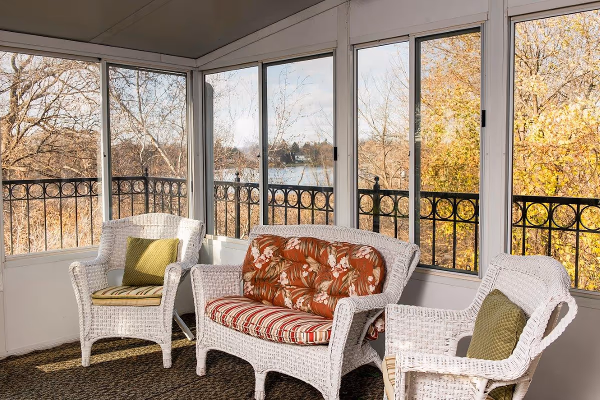 A sunroom with white wicker furniture including two armchairs and a loveseat, each with cushions. The room has large windows showing a view of trees with autumn foliage and a body of water in the background.