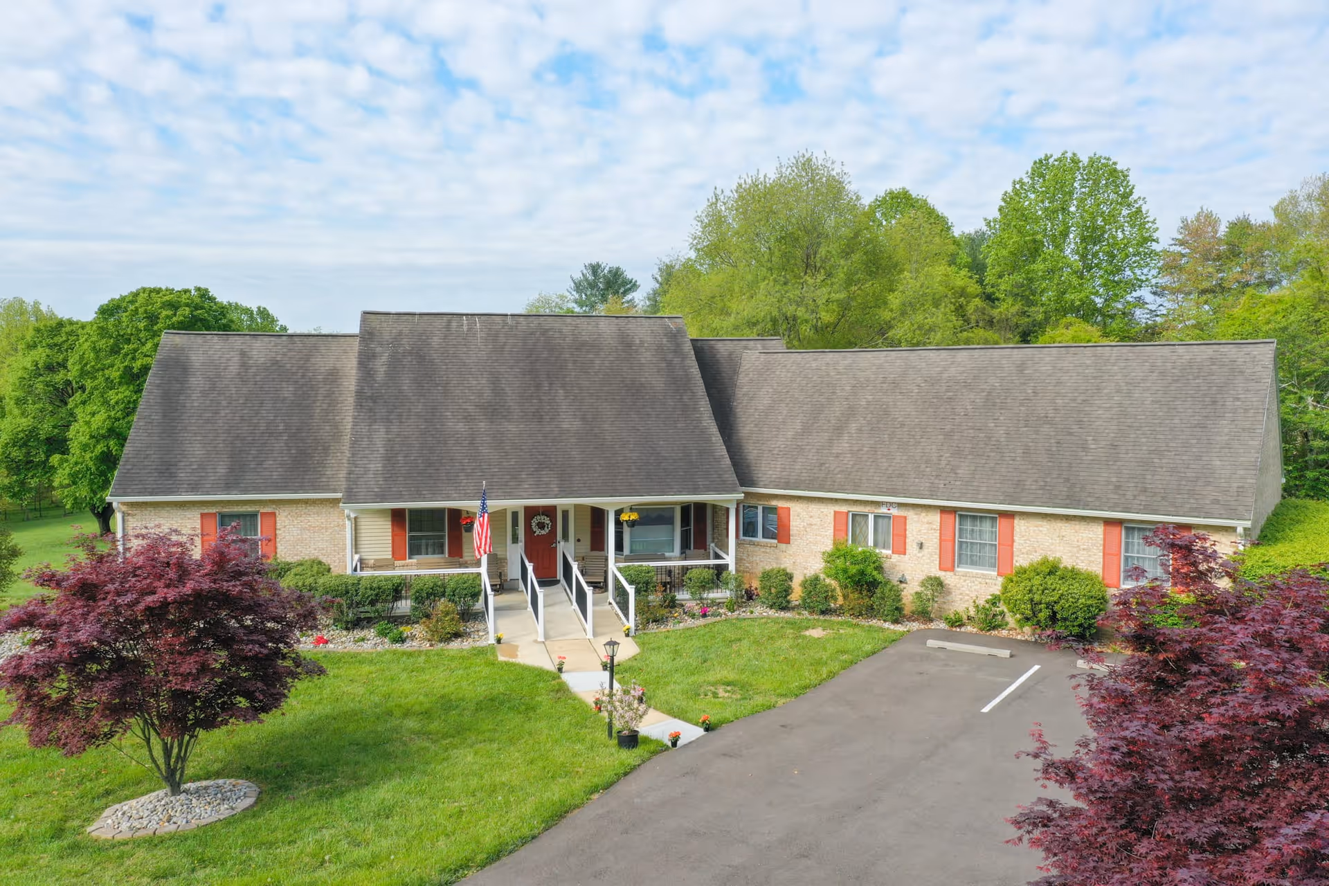 Front exterior view of a single-story residential building with a large sloped roof, beige brick walls, red shutters, and a central entrance with a ramp and steps. The building is surrounded by green grass, small trees, and shrubs under a partly cloudy sky.