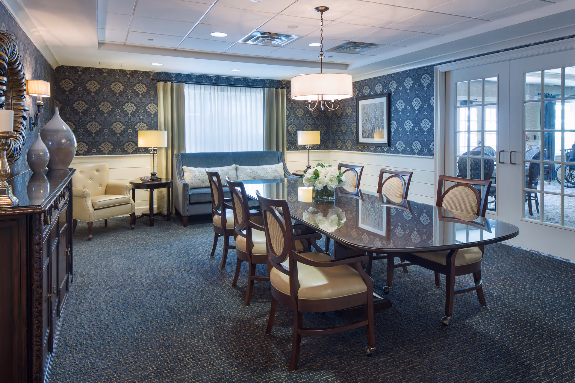 A formal dining room with a long polished wooden table surrounded by eight cushioned chairs. The room features patterned wallpaper, a large window with curtains, two table lamps, a hanging ceiling light, a sideboard with decorative vases, and a blue upholstered sofa with cushions. French doors lead to another room.