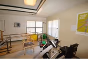 A small exercise room with various fitness equipment including a stationary bike, handrails for walking support, a chair, and exercise balls. The room has large windows with blinds, light-colored walls, and a ceiling light. A poster is visible on the wall.