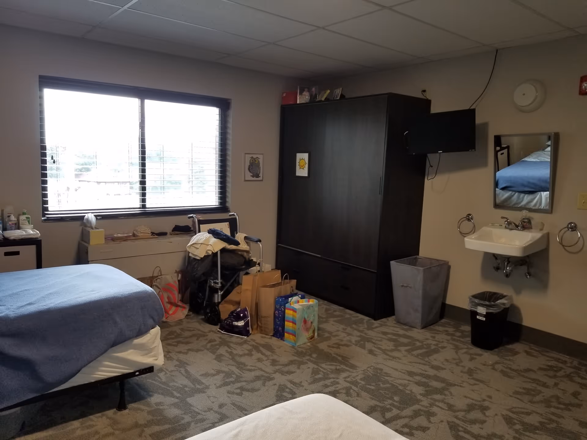 A senior living resident room with beds, a large window, a dark wardrobe, a wall-mounted TV, and a sink.