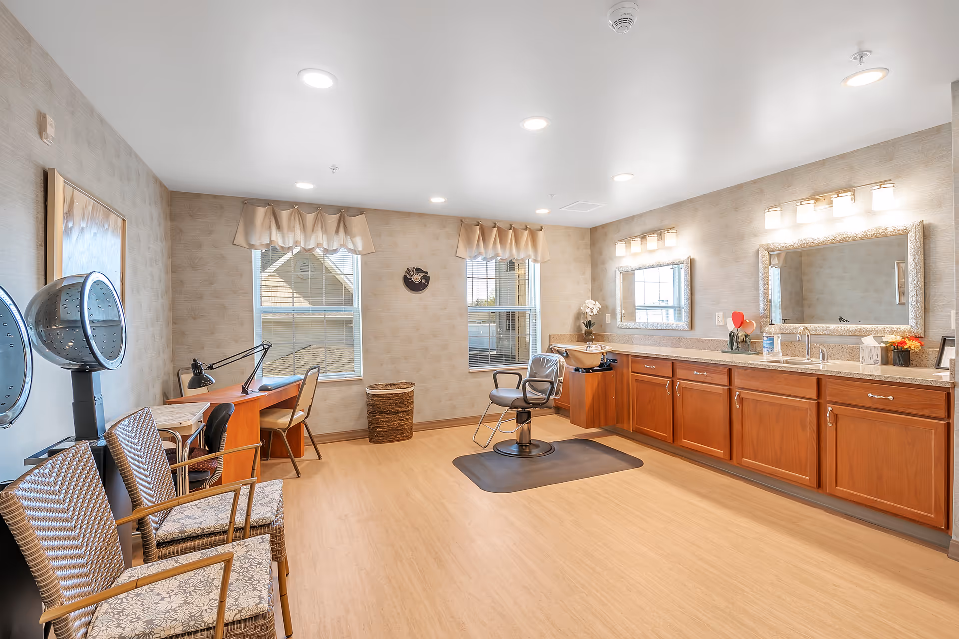 A bright and spacious salon room with two hair drying chairs on the left, a desk with a lamp and chair near two windows with beige valances, a single salon chair on a black mat in the center, and a long wooden cabinet with two large mirrors and sinks on the right wall.