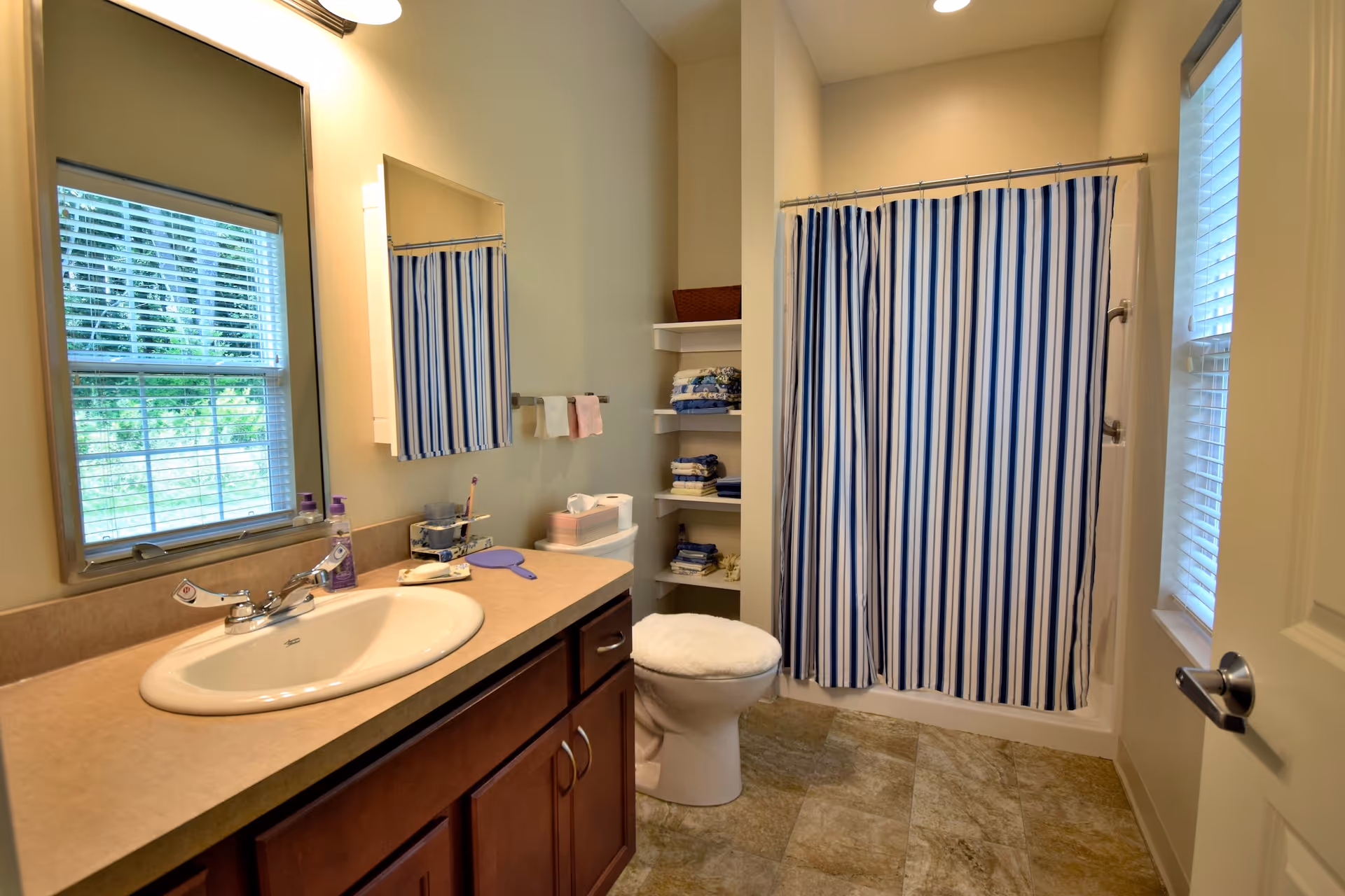 Bathroom with a sink and countertop on the left, a mirror above the sink, a window with blinds reflecting in the mirror, a toilet with a cushioned seat cover, shelves with folded towels, and a shower with a blue and white striped curtain. The floor is tiled and there is a door on the right side.
