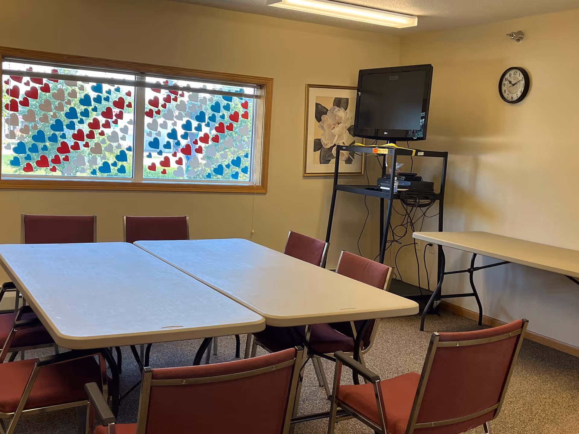 A small meeting or activity room with two rectangular tables pushed together surrounded by red cushioned chairs. A window decorated with red, white, and blue heart-shaped stickers is on the left wall. On the right wall, there is a clock showing the time as 3:15, a framed floral picture, and a black TV mounted on a metal stand with electronic devices and cables underneath. Another folding table is against the right wall.