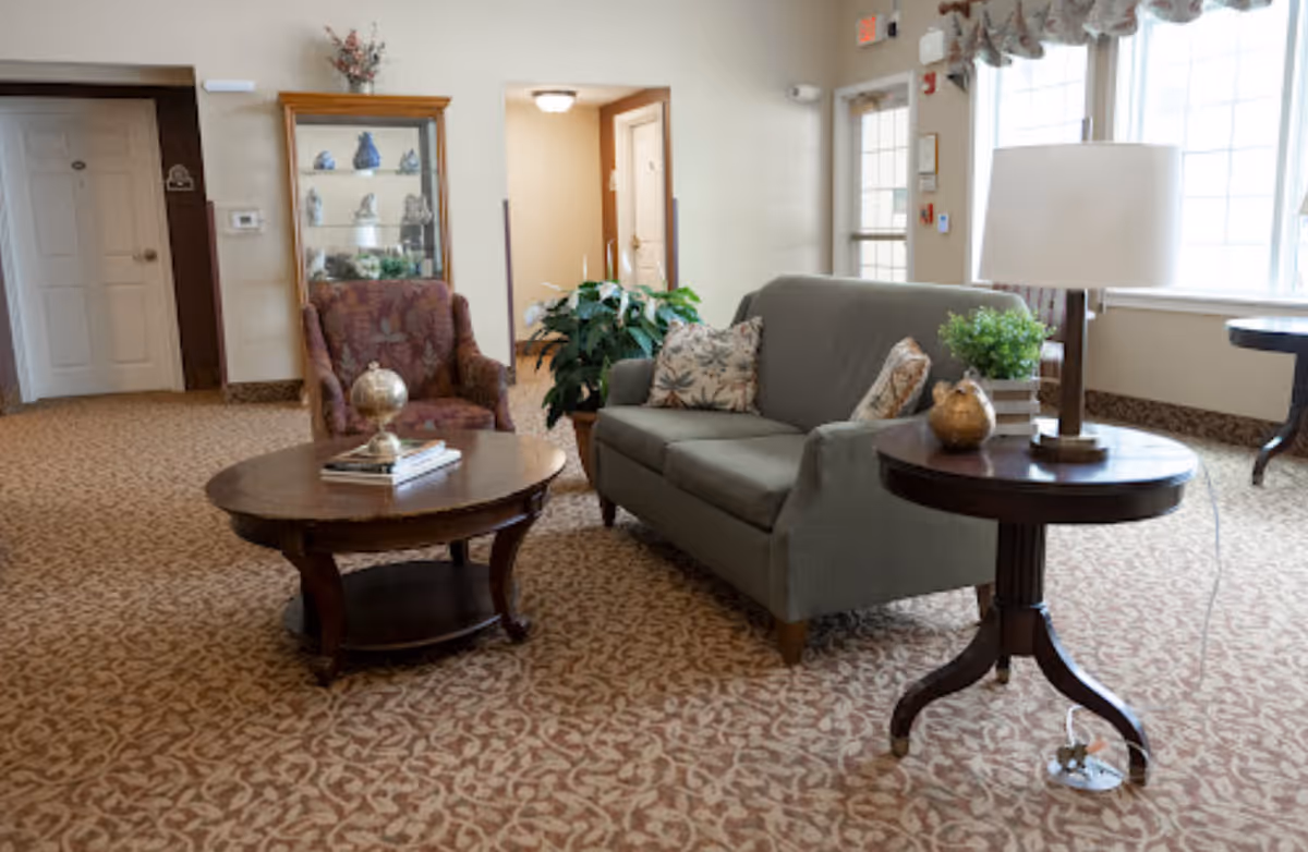 A cozy senior living facility common area with a patterned carpet, a gray sofa with decorative pillows, a floral armchair, a round wooden coffee table with books and a decorative globe, a side table with a lamp and a small plant, and a glass display cabinet with decorative items. Large windows allow natural light to fill the room.