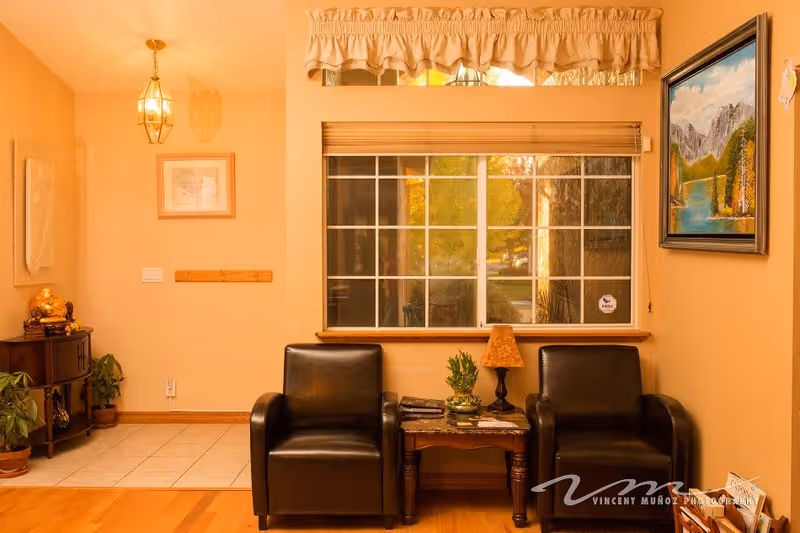 A cozy interior seating area with two black leather armchairs separated by a small wooden table holding a lamp and a plant. Behind the chairs is a large window with blinds and a valance. The walls are painted a warm beige color and decorated with framed artwork. To the left, there is a small corner table with a golden Buddha statue and some plants on the floor. The floor is a combination of wood and tile.