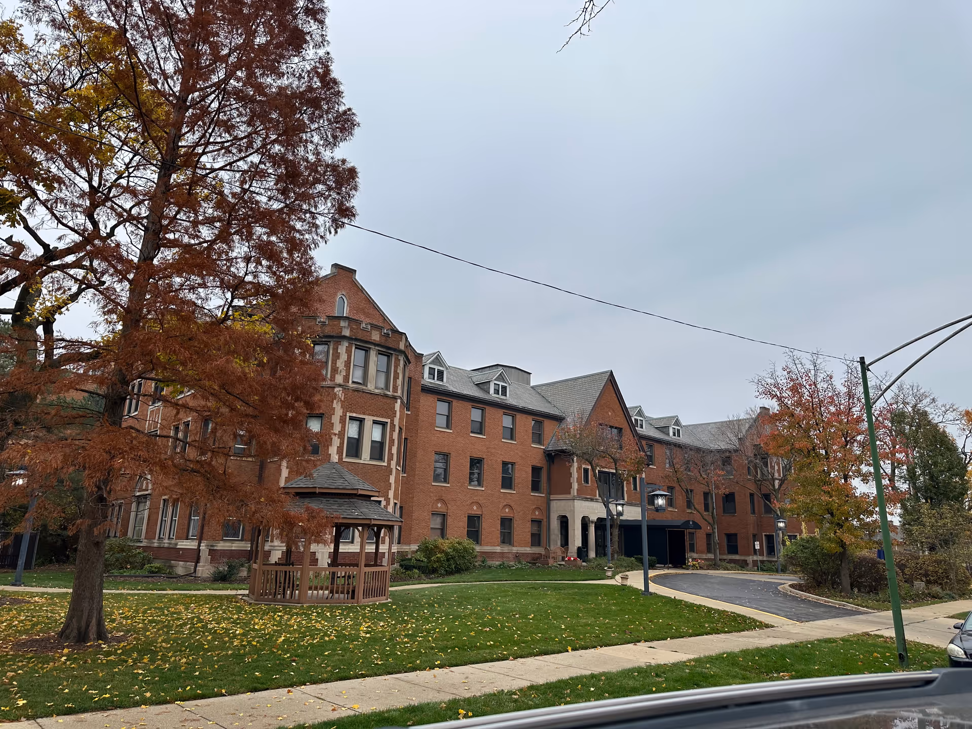 Brick multi-story residential building with a small wooden gazebo on a grassy lawn and autumn trees under a gray sky.