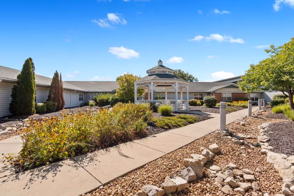 Outdoor courtyard area of EverTrue Breeze Park featuring a white gazebo surrounded by landscaped garden beds with shrubs and trees, a paved walkway, and a clear blue sky.