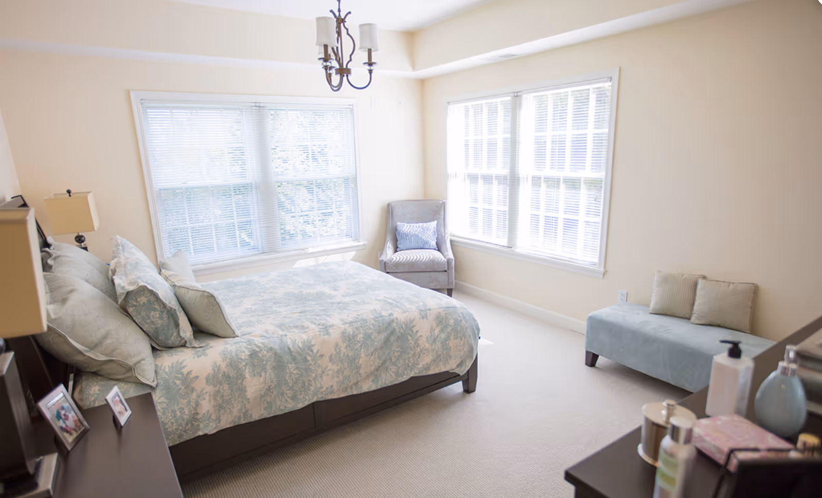 Sunlit bedroom with a bed covered in patterned bedding, large windows, an armchair, and a small bench.