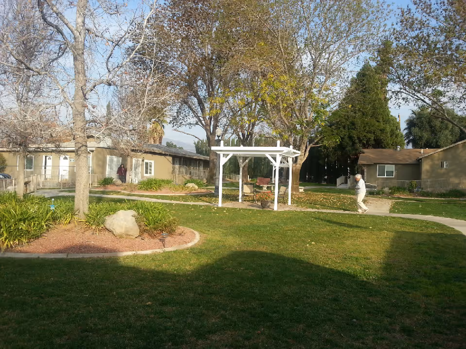 Sunny courtyard with lawn, trees, a white pergola and single-story residential buildings, with a person walking on a path.