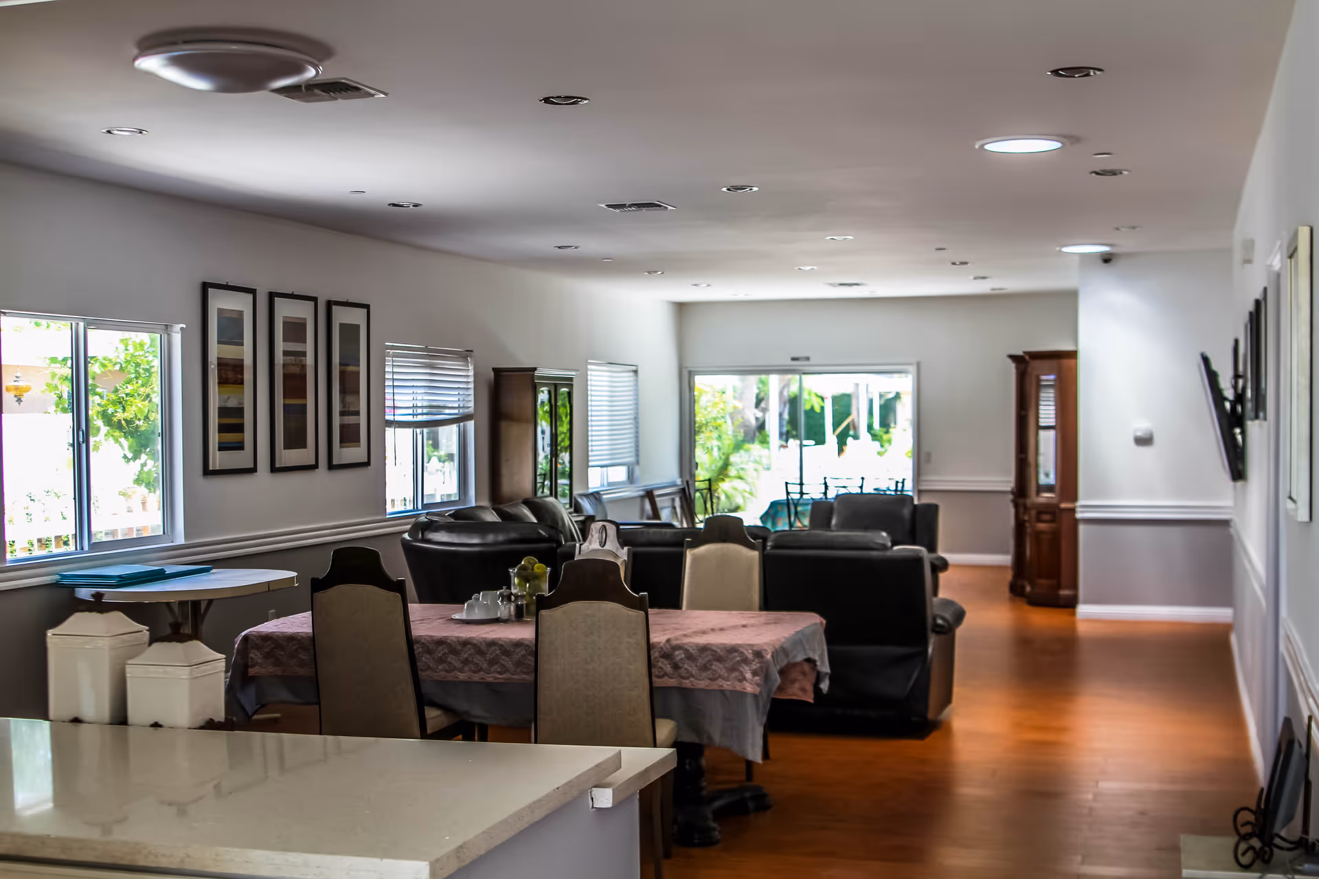 Interior view of a senior living facility common area with a dining table covered with a pink tablecloth and chairs, leather sofas arranged in a seating area, framed artwork on the wall, windows with blinds, and a glass door leading to an outdoor space.
