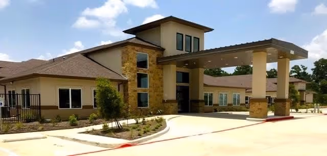 Exterior view of a modern single-story building with stone and beige stucco walls, large windows, and a covered entrance with pillars. The building is surrounded by a paved driveway and landscaped with small shrubs and trees under a partly cloudy sky.