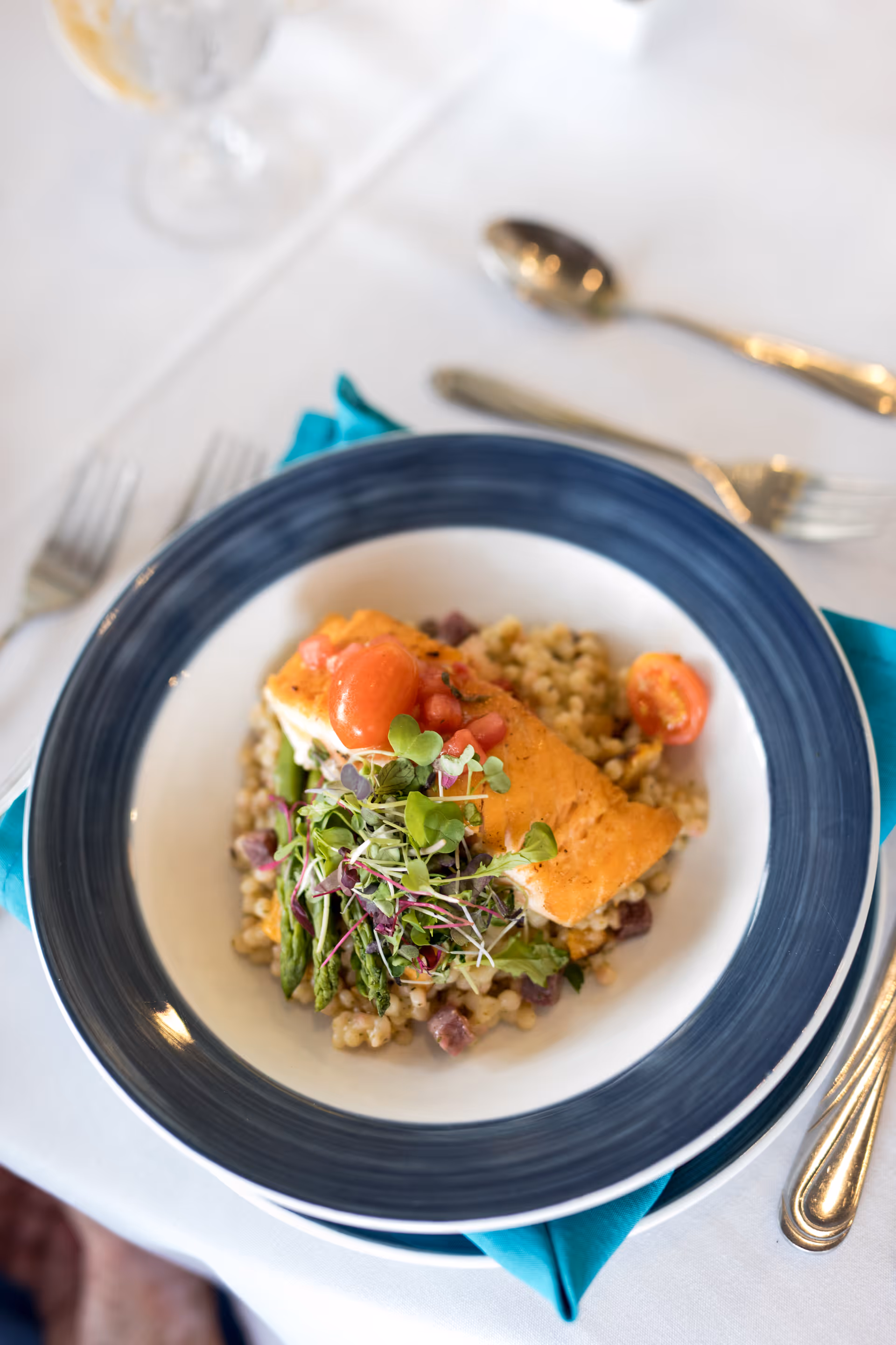 A plated salmon entrée with grains, asparagus, microgreens and cherry tomatoes on a blue-rimmed bowl set on a white tablecloth with silverware.
