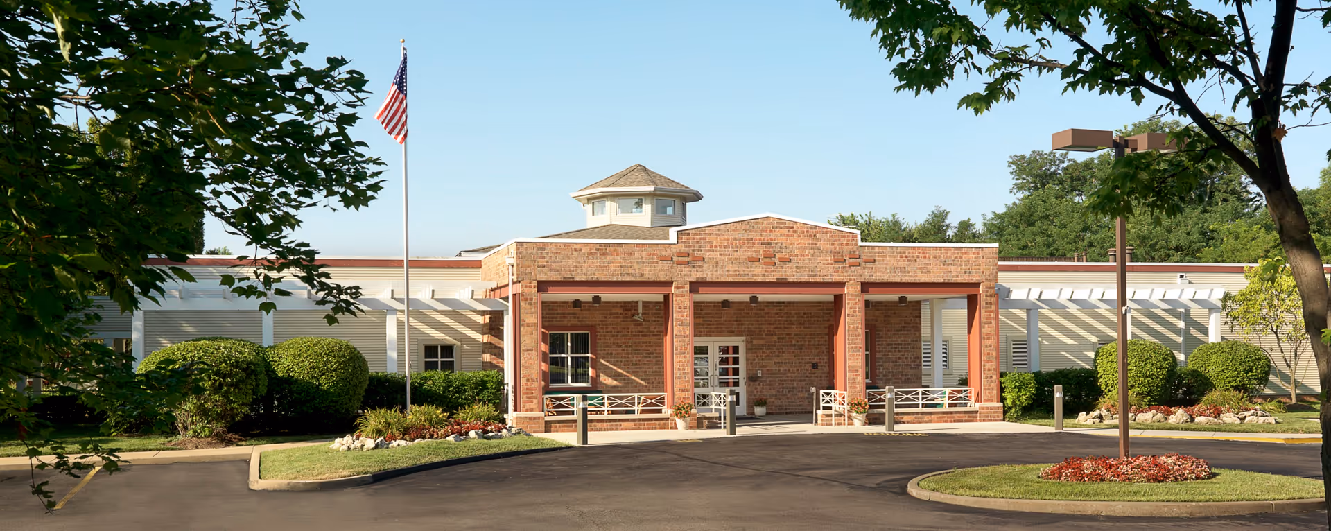 Front exterior view of a single-story brick building with a covered entrance supported by columns, surrounded by well-maintained landscaping including bushes and trees, with an American flag on a flagpole in front.
