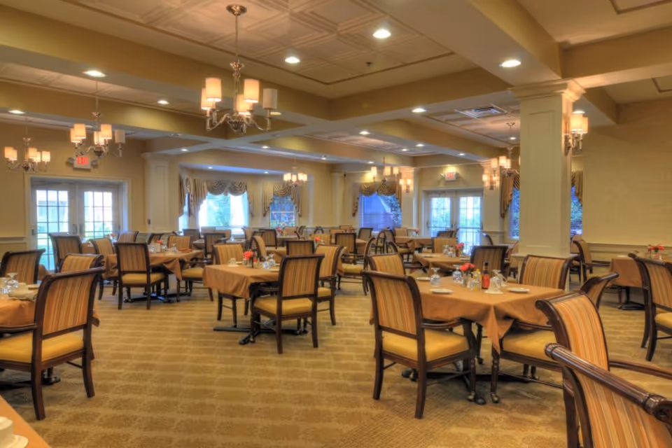 A spacious dining room with multiple tables covered in brown tablecloths, each set with glassware, plates, and small flower arrangements. The room features striped upholstered chairs, chandeliers hanging from a coffered ceiling, large windows with draped curtains, and soft lighting creating a warm atmosphere.