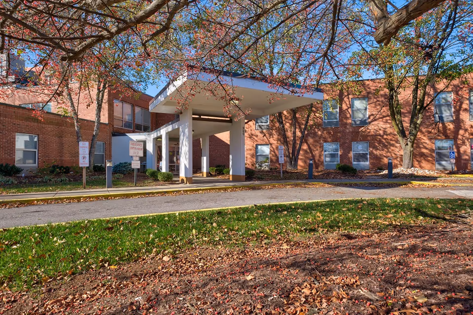 Exterior view of a brick building with a covered entrance supported by white pillars. Trees with autumn leaves frame the scene, and there are signs indicating no parking and ambulance entry. The sky is clear and blue.