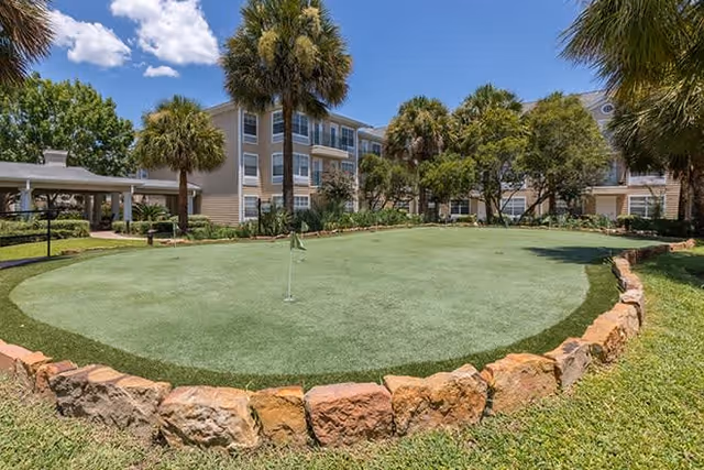 Outdoor putting green surrounded by a low stone border with palm trees and bushes around it, set in front of a multi-story residential building under a blue sky with some clouds.