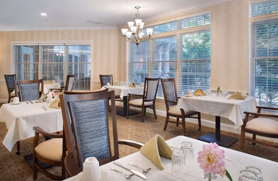 Bright dining room with multiple set tables and wooden chairs, large windows, and a central chandelier.
