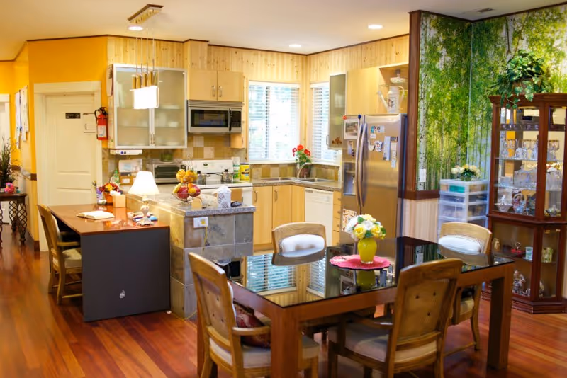 Open-plan kitchen and dining area with a glass-top table and wooden chairs, a central island, light wood cabinets and a stainless steel refrigerator.