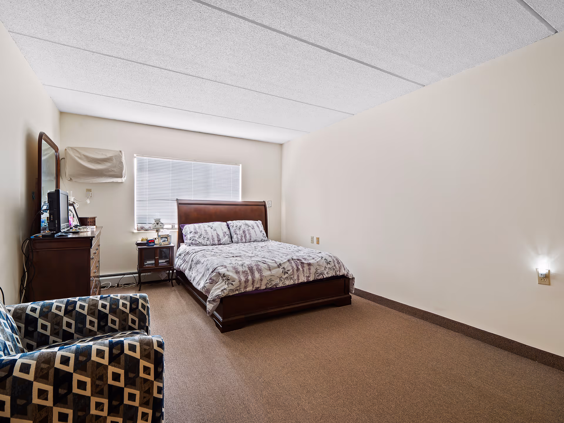 Bedroom with a wooden bed and patterned bedding, a dresser with mirror, a bedside table, and a patterned armchair under a window with blinds.