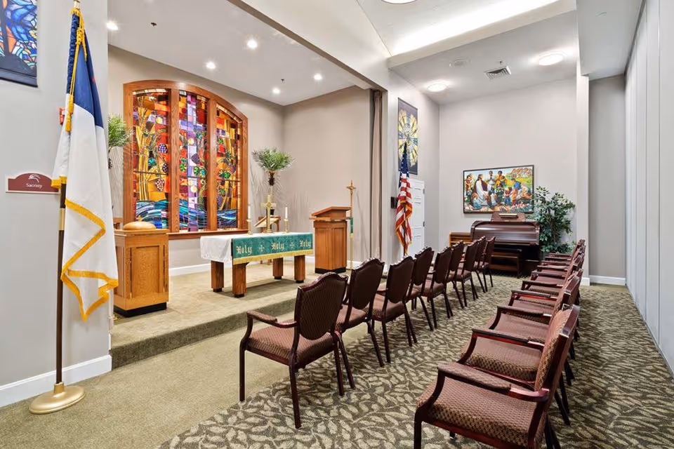 Interior view of a small chapel or worship room with rows of chairs facing an altar covered with a green cloth that reads 'Holy Holy Holy'. There is a stained glass window behind the altar, a wooden podium, two flags on stands, a piano, and religious artwork on the walls.