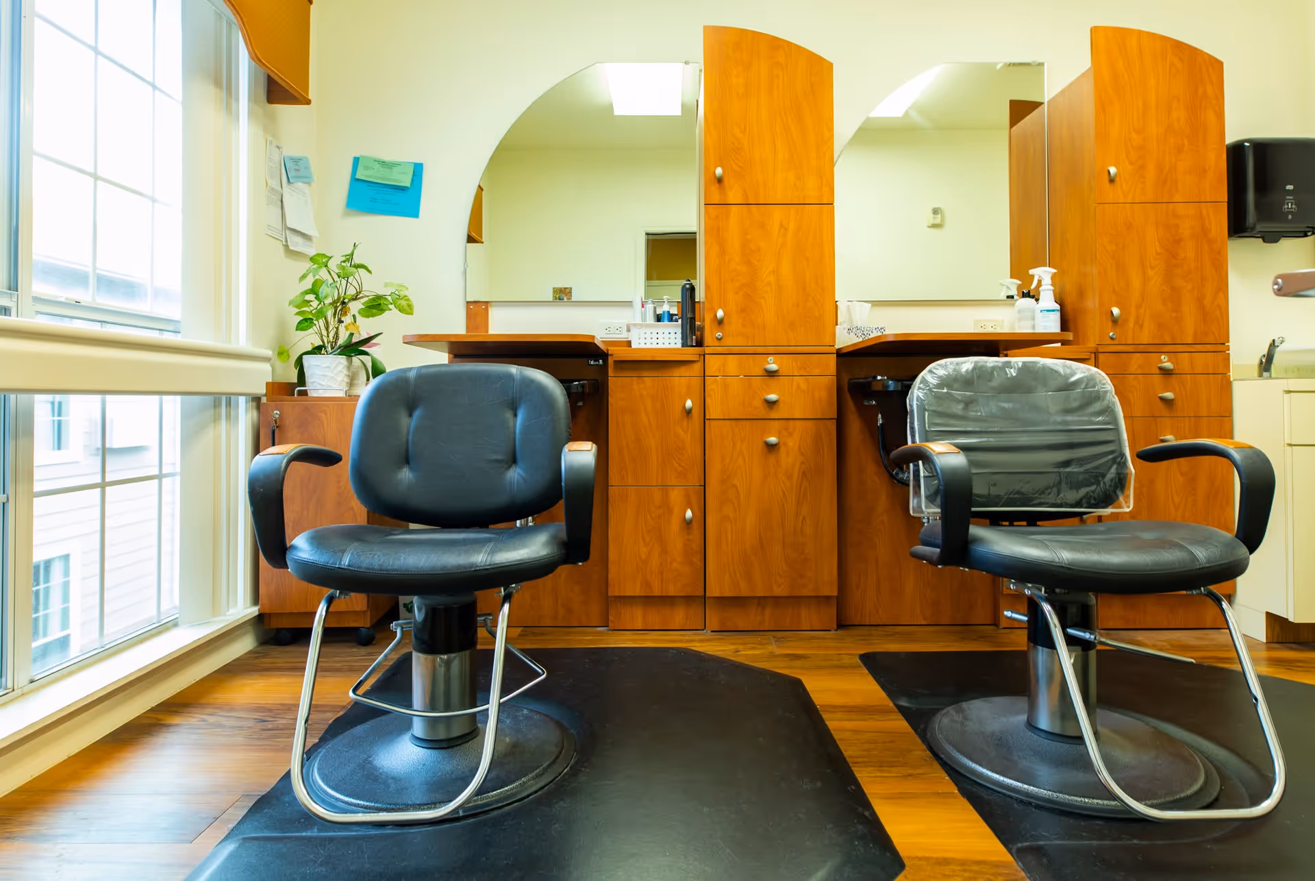 Interior view of a hair salon area in a senior living facility with two black salon chairs in front of wooden cabinets and mirrors. There is a large window on the left side allowing natural light to brighten the room, and various hair care products and a plant are visible on the counter and cabinet.