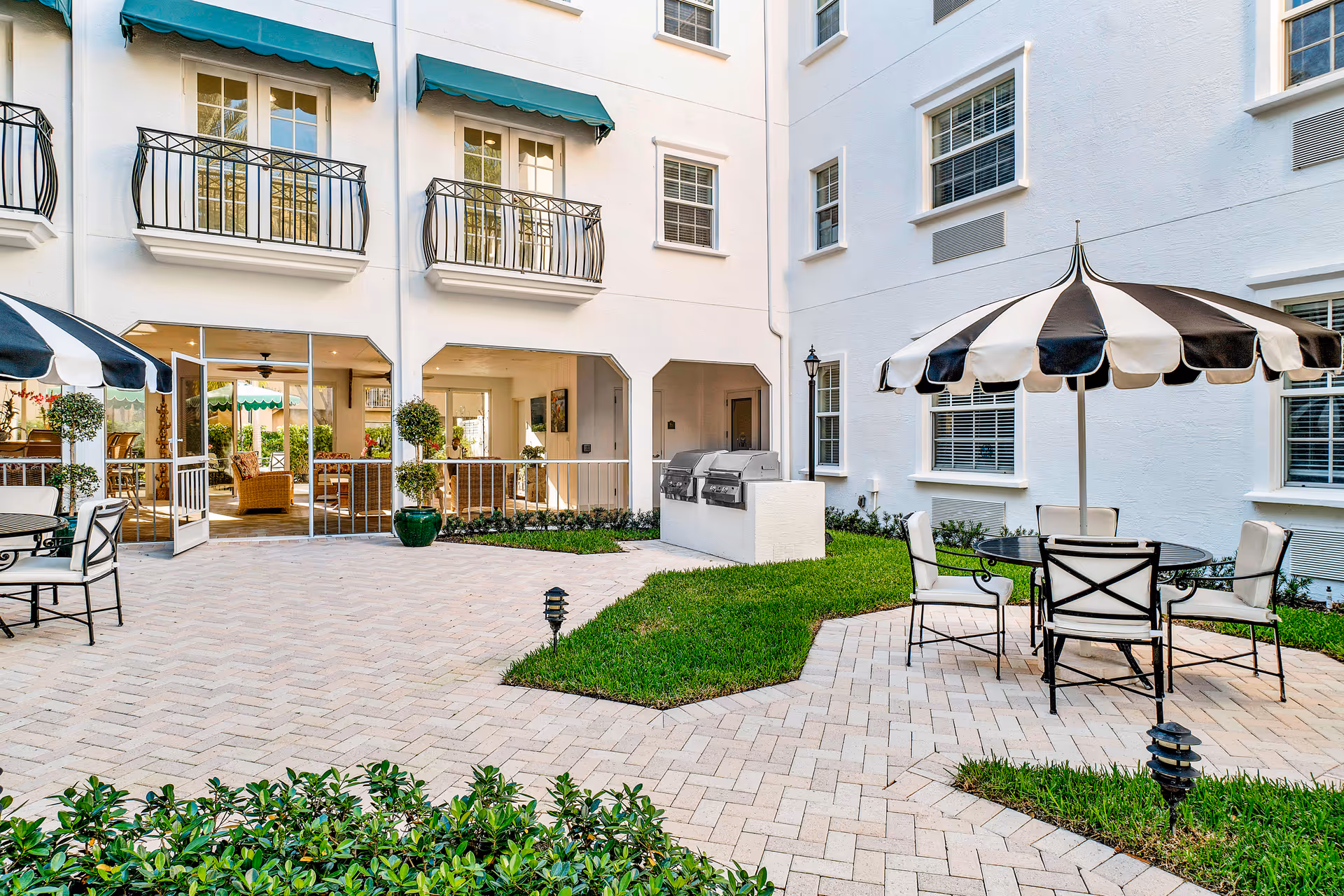 Courtyard patio with tables, striped umbrellas, seating, greenery and a built-in grill beside a white multi-story building.
