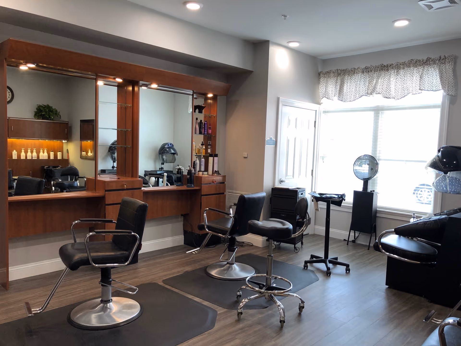 Interior view of a hair salon area with multiple black salon chairs, large mirrors with wooden frames, hair care products on shelves, and hair drying stations near a large window with patterned curtains.