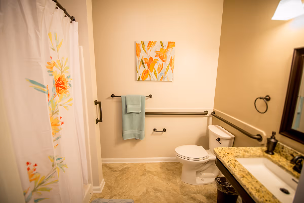A clean and well-lit bathroom featuring a white toilet, a granite countertop with a sink, a mirror, and a shower with a floral-patterned curtain. There are safety grab bars installed near the toilet and along the wall, a towel rack with a light blue towel, and a colorful floral painting on the wall.