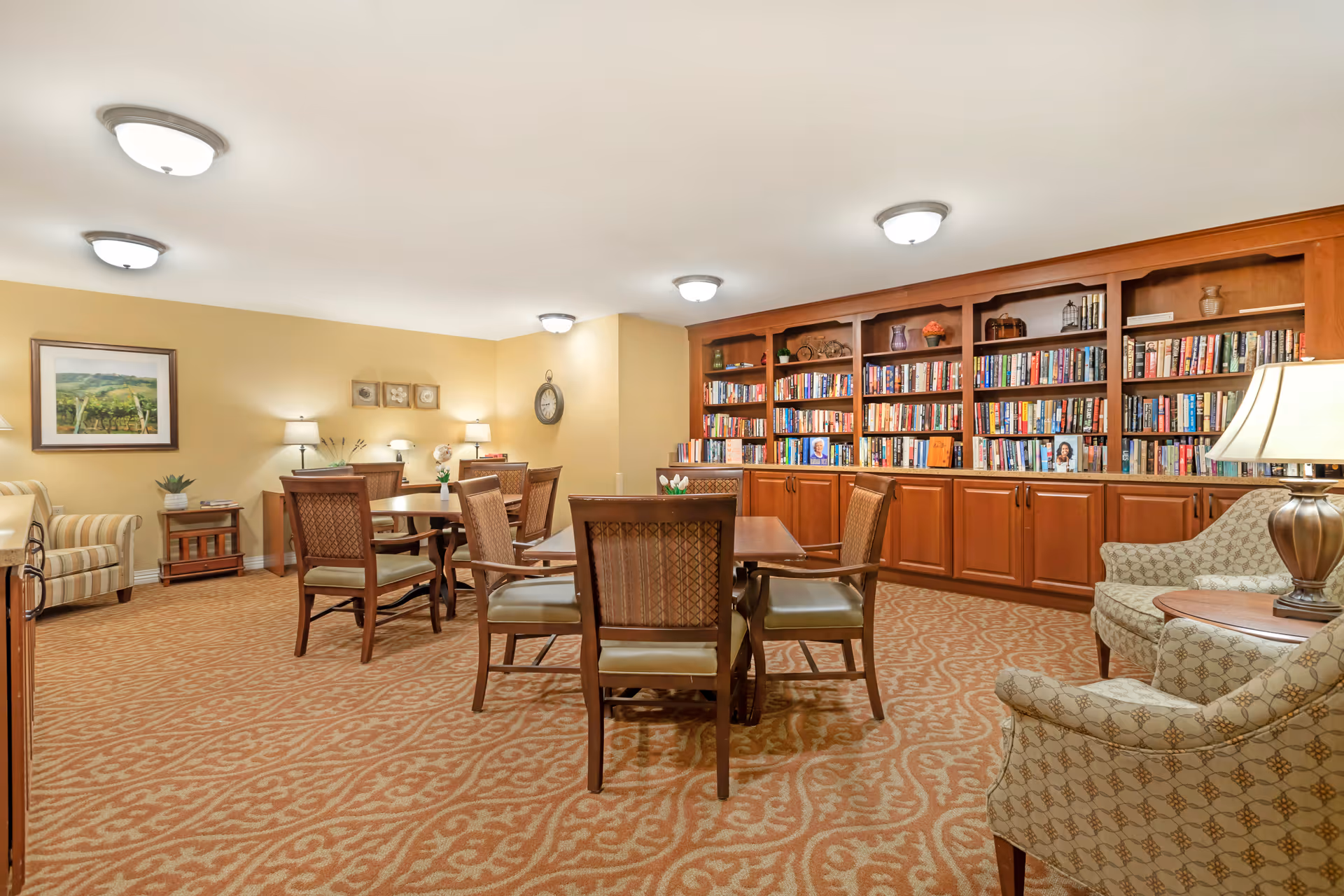 A cozy library room with wooden bookshelves filled with books along the back wall. There are several wooden tables with cushioned chairs arranged around the room. The carpet has a patterned design, and the walls are painted a warm beige color. The room is well-lit with ceiling lights and table lamps, and there are framed pictures and a clock on the walls.