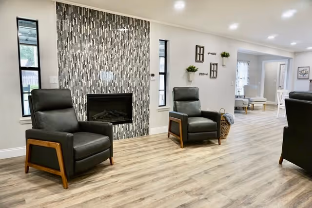 Bright communal living room with two dark leather armchairs facing a modern fireplace set in a vertical mosaic tile wall and an open dining area beyond.